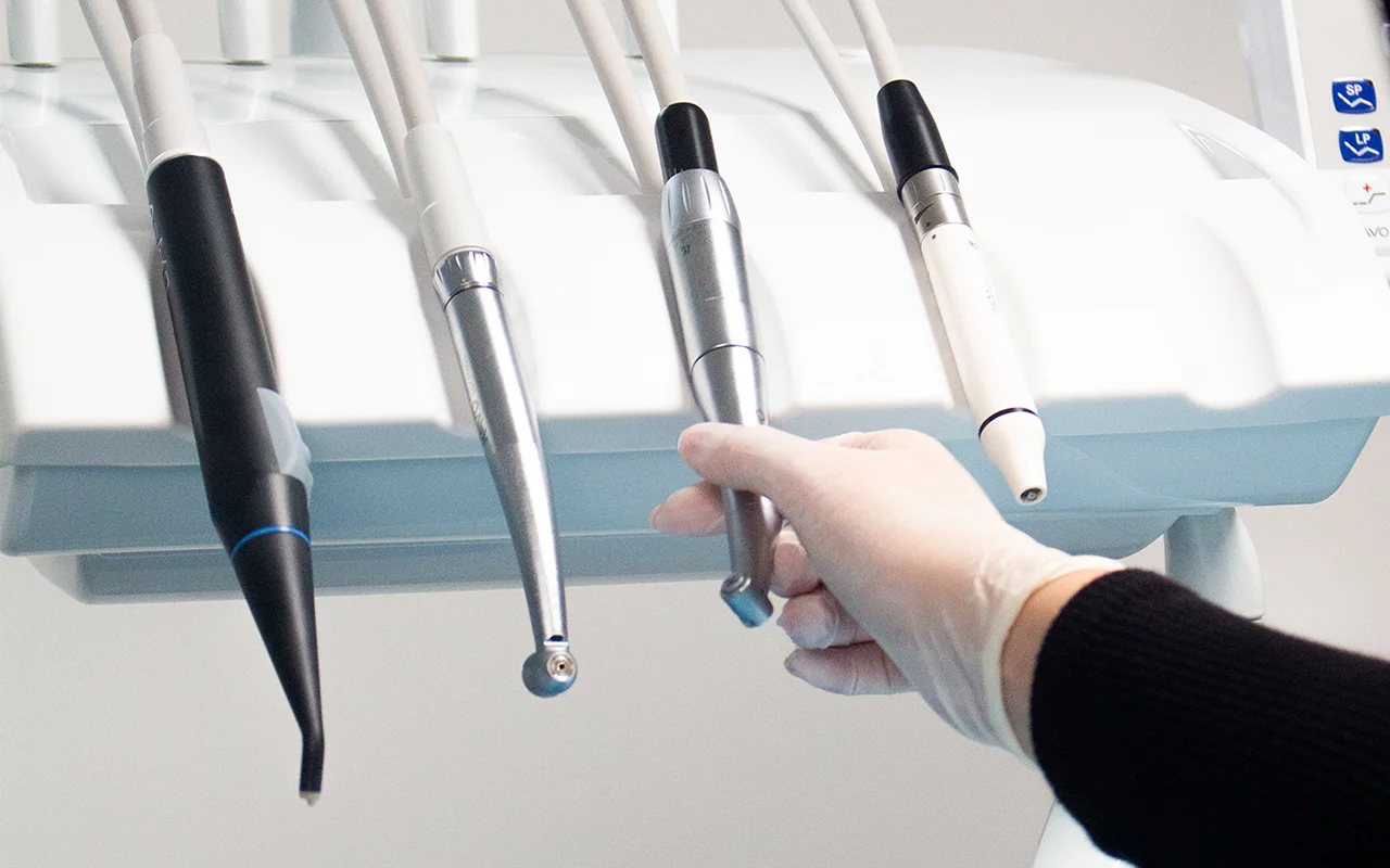 Mão com luva a segurar peça de equipamento dentário com sugador e outros instrumentos ao fundo.