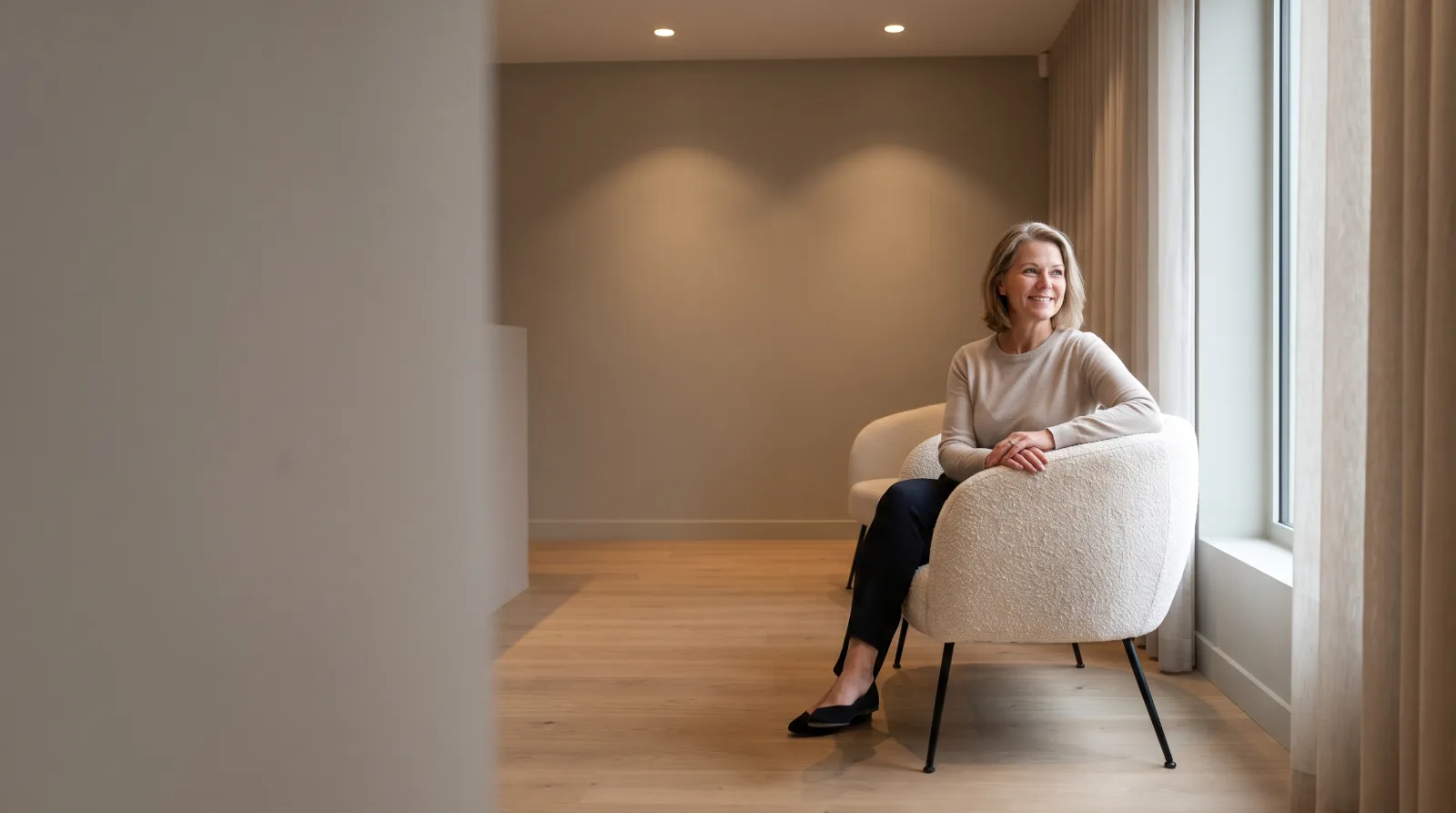Mulher madura a sorrir de forma serena, sentada numa poltrona creme num ambiente claro e acolhedor.